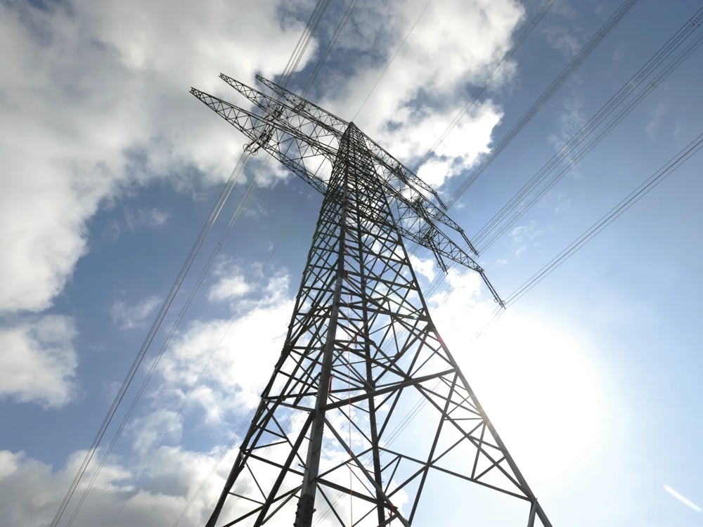 Ein Strommast ragt in den blauen Himmel, umgeben von weißen Wolken.