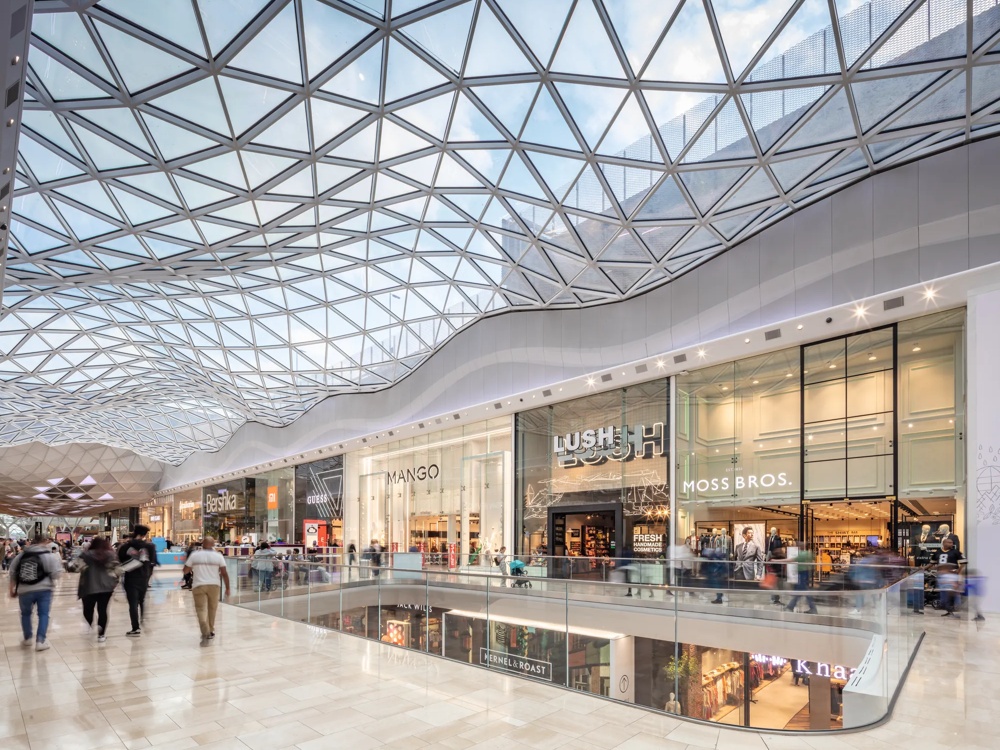 Eine moderne Einkaufspassage mit gläsernem Dach, Geschäften und Menschen, die entlang eines Geländers schlendern.