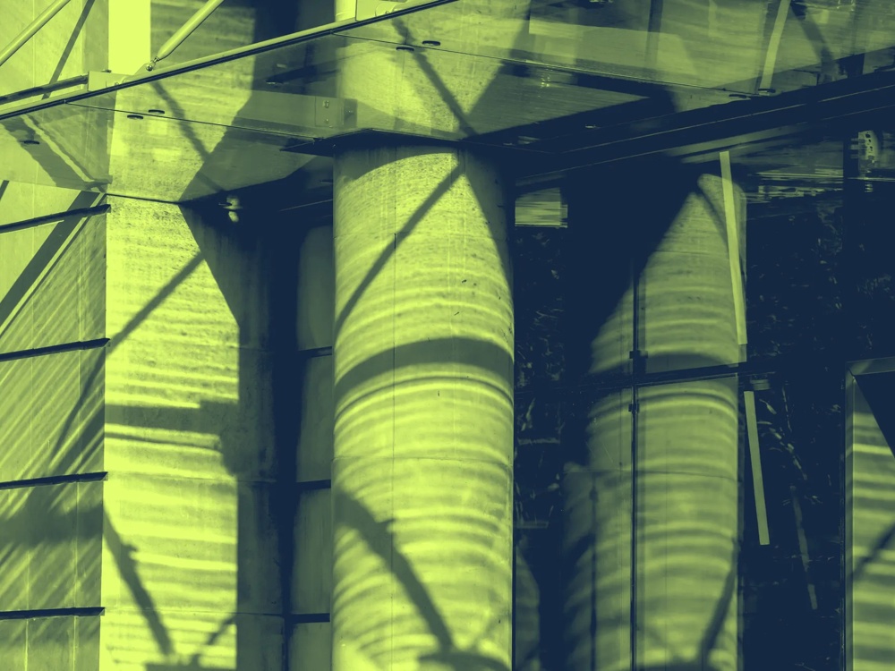 Schattenmuster auf Glasfassade mit Betonpfeilern und Metallstrukturen in gelbgrünem Licht.