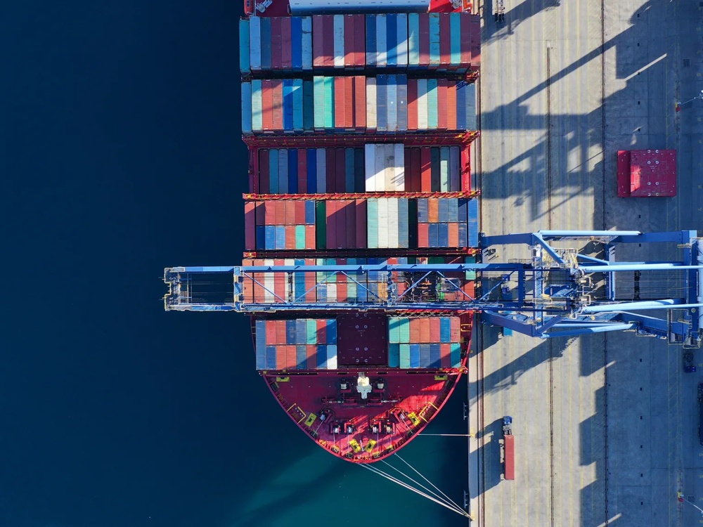 Ein Containerschiff mit bunten Containern liegt an einem Hafen, neben einem großen blauen Kran.