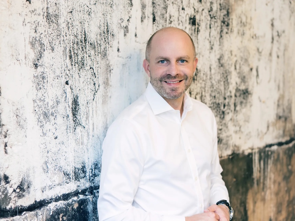 A man in a white shirt leaning on a structured, weathered wall with a smile.