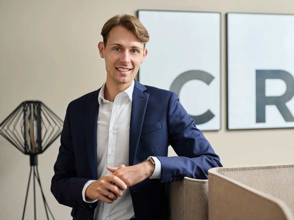 Leon Christmann ist Berater Young Professionals. Leon Christmann steht vor einem hellen Hintergrund, hinter ihm sind die gerahmten Initialen der Commerz Real und eine schwarze Stehlampe zu sehen.