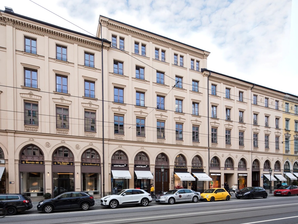 Elegantes, mehrstöckiges Gebäude mit Arkaden und Schaufenstern, davor parkende Autos und Passanten.