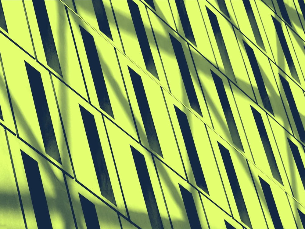 Gelbe Wand mit diagonalen, schmalen, dunklen Fenstern im geometrischen Muster, Schatten fallen schräg über die Oberfläche.