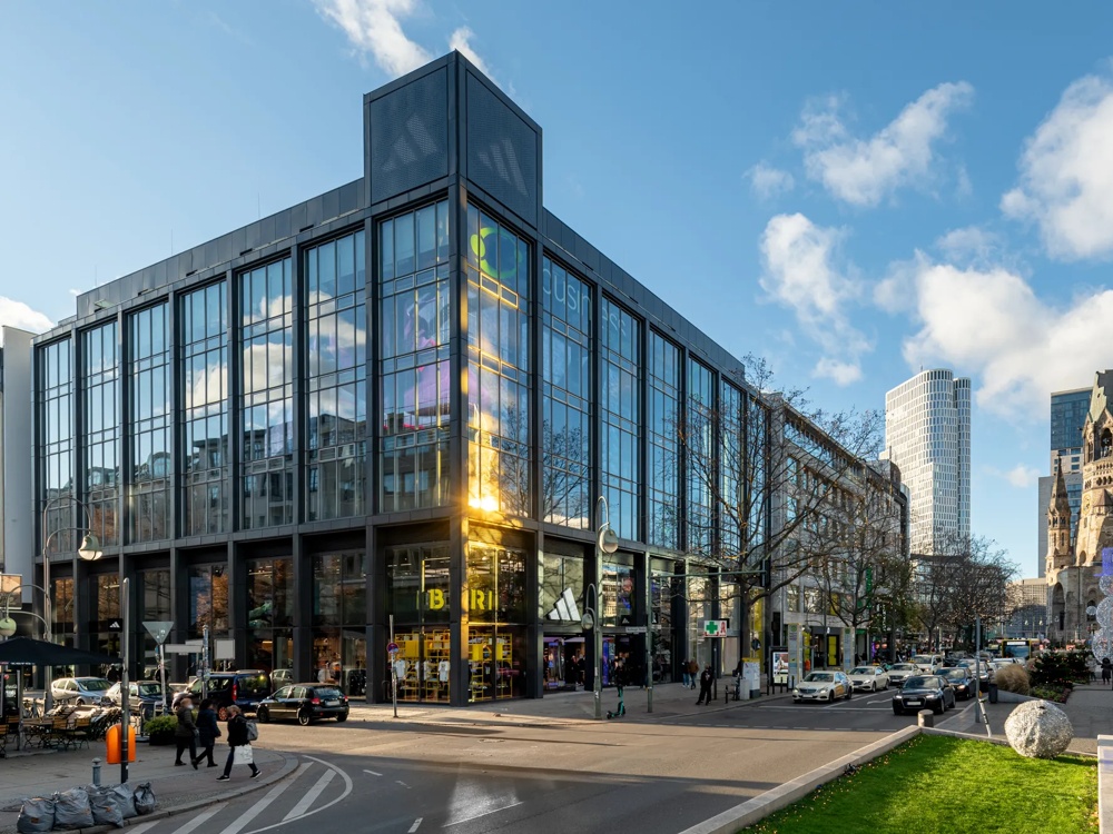 Moderne Glasfassade eines Gebäudes bei Sonnenschein, Menschen und Autos auf der Straße, blauer Himmel mit Wolken.