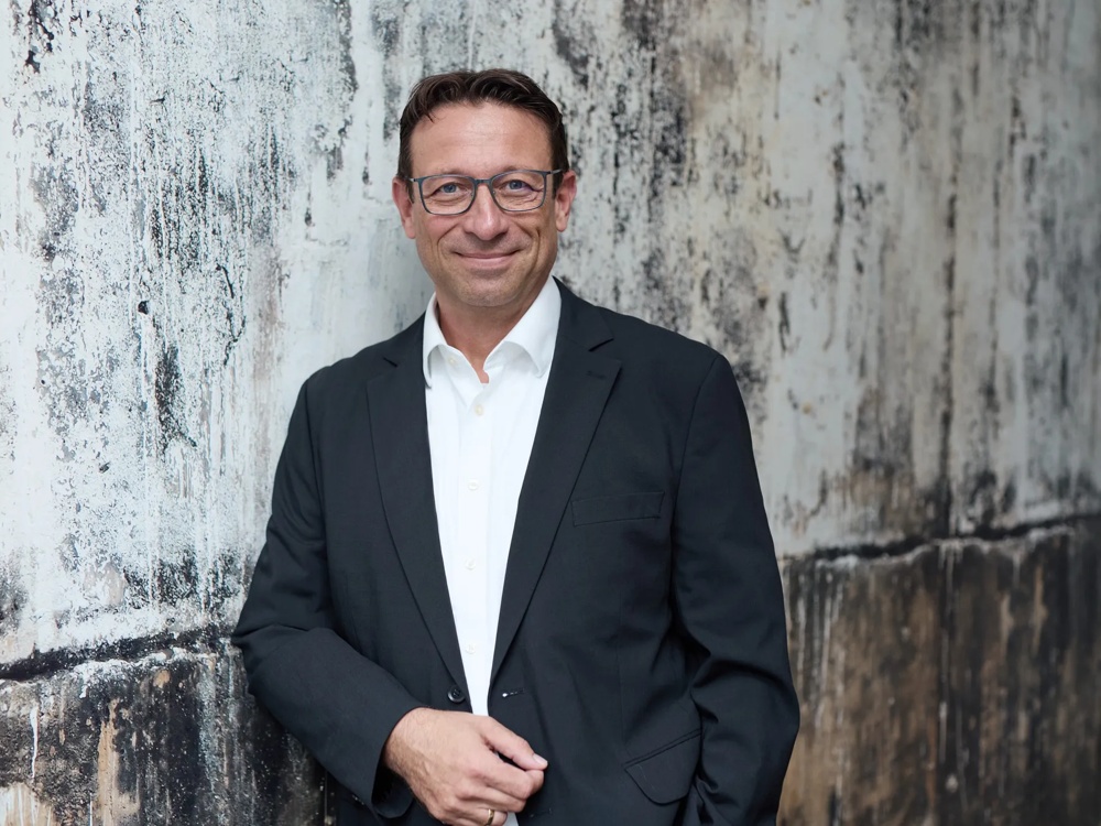 Ein Mann in Anzug und Brille lehnt lächelnd an einer strukturierten, verwitterten Wand.