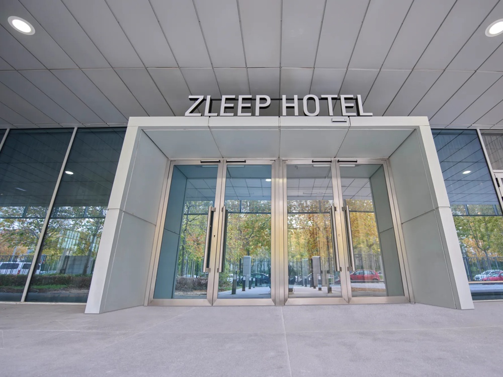 Eingang des Zleep Hotels mit Glasdrehtüren und reflektierenden Fenstern, Bäume im Hintergrund.