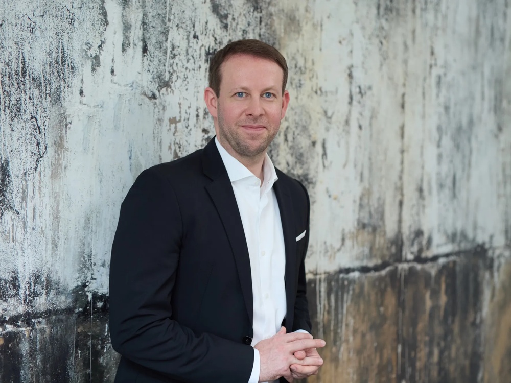 The Commerz Real employee Christian Rockel, Head of Sales Private & Business Clients NOM for Sales Private and Business Clients NOM, is represented. He is standing in front of a textured concrete wall, wearing a black blazer with a white shirt and smiling lightly into the camera.
