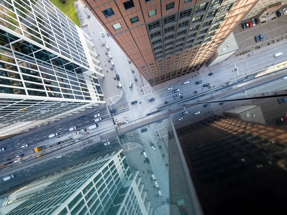 Blick von oben auf eine belebte Kreuzung zwischen hohen Gebäuden, mit Autos auf der Straße und Spiegelungen in den Glasfassaden.