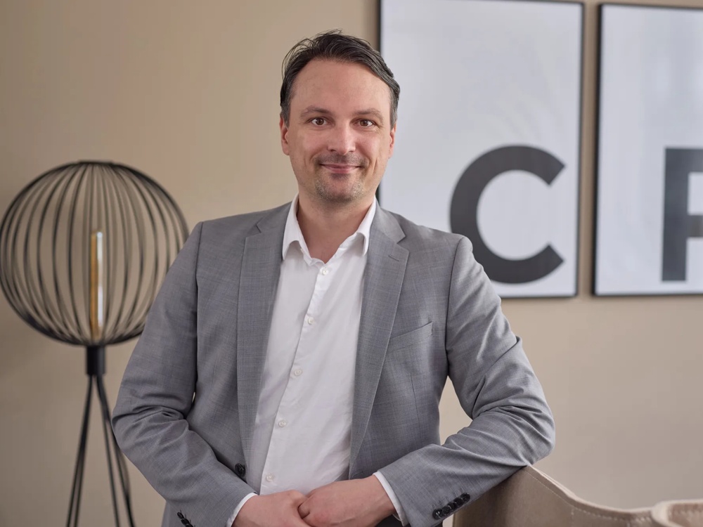 Ein Mann im grauen Anzug steht lächelnd vor zwei gerahmten Buchstaben an der Wand.