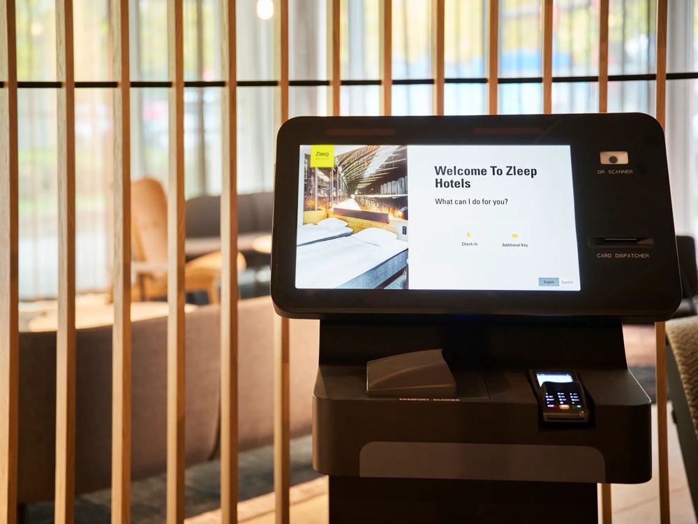 A check-in kiosk in a hotel with the screen text 'Welcome to Zleep Hotels’ in front of wooden battens.