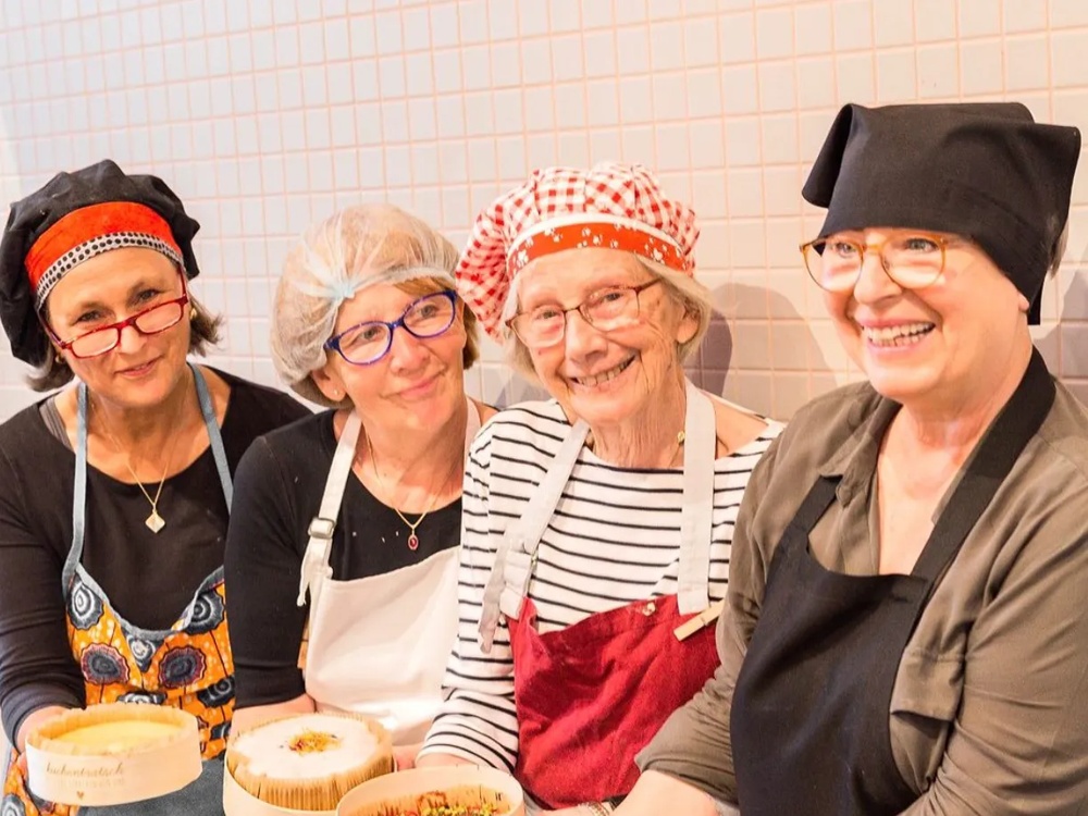 Vier lächelnde Frauen in Schürzen und Kochmützen halten Kuchen in runden Holzformen.