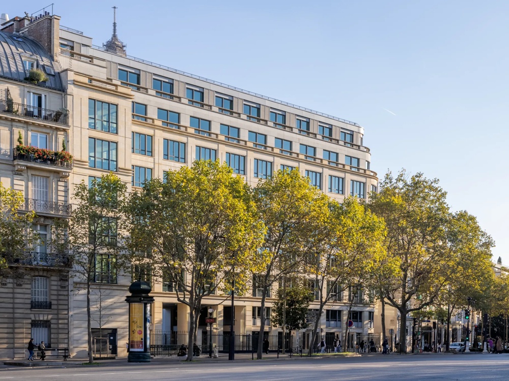 Ein großes, modernes Gebäude mit vielen Fenstern, umgeben von Bäumen an einer belebten Straße bei Tageslicht.