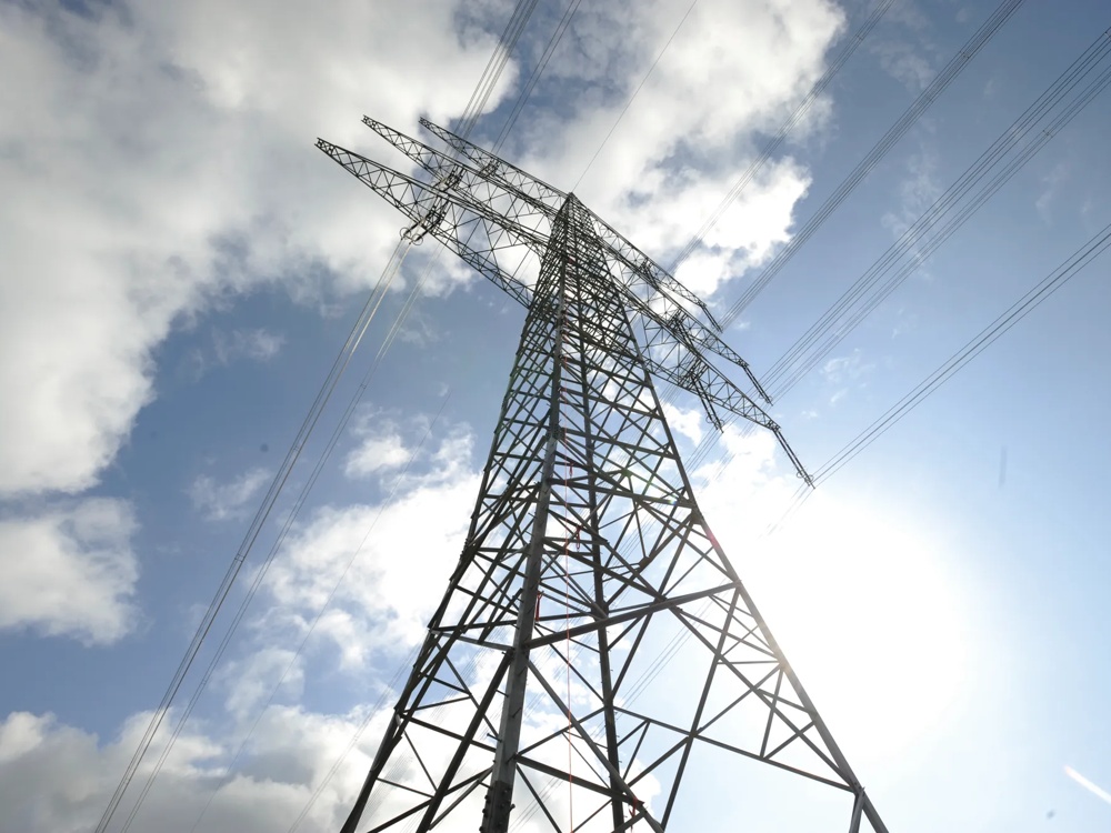 Strommast ragt in den blauen Himmel, umgeben von Wolken und Sonnenlicht.