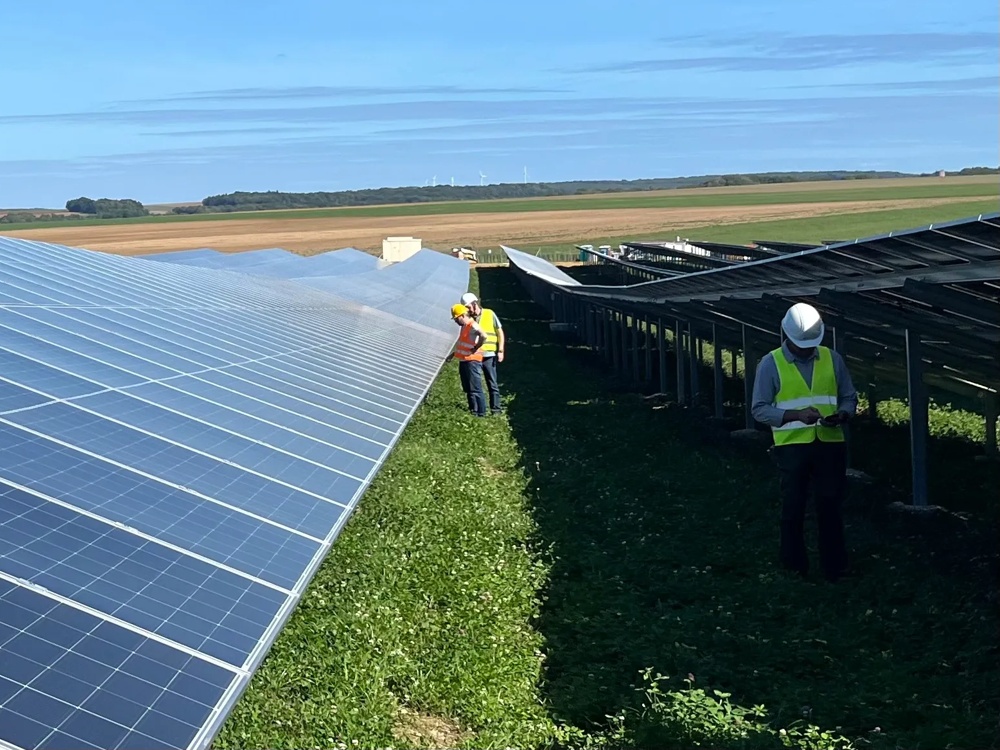 Zwei Personen in Warnwesten inspizieren Solarpanels auf einem grünen Feld unter blauem Himmel.