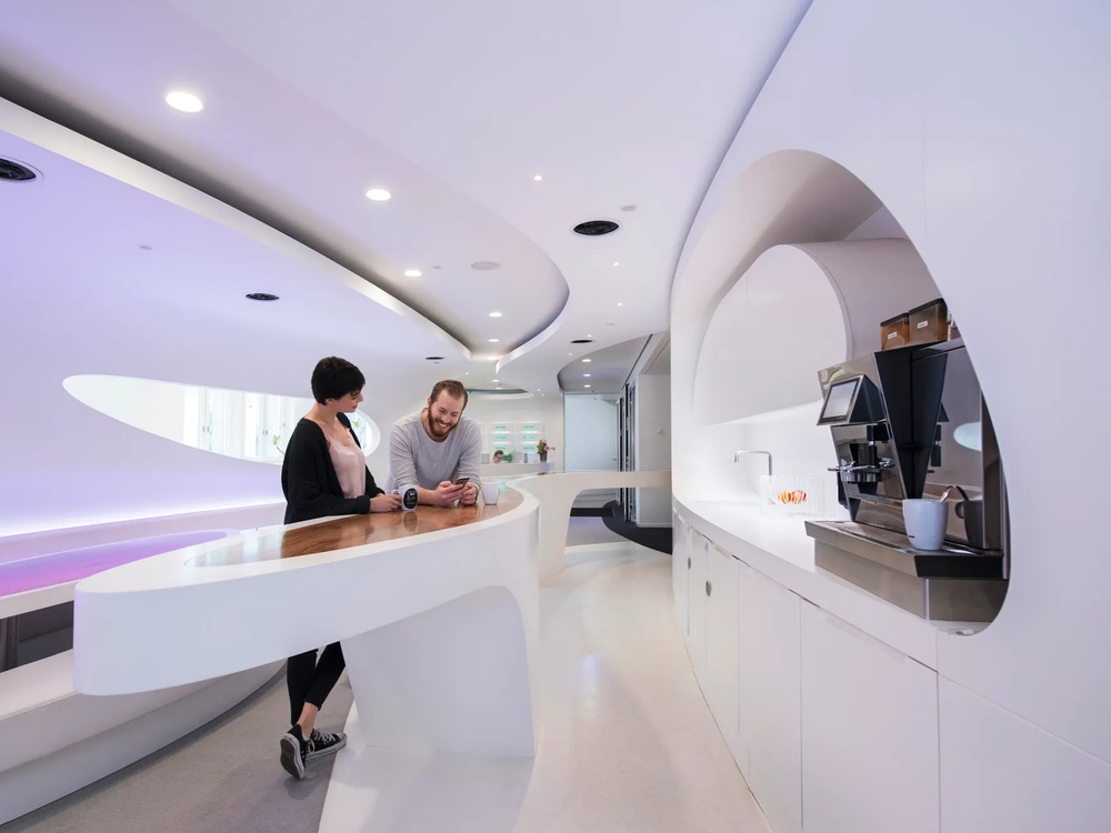 Futuristisches Büro mit geschwungenen weißen Wänden, Holztresen, zwei Personen und Kaffeemaschine.