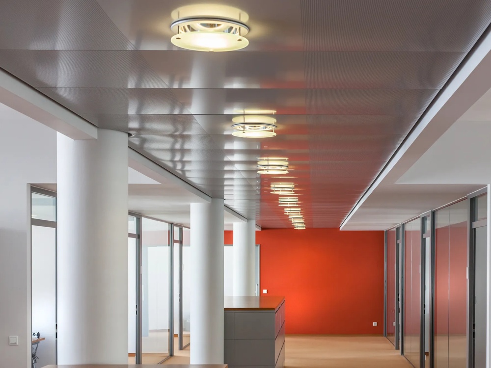 Moderner Büroflur mit roter Wand, gläsernen Büros, weißen Säulen und runder Deckenbeleuchtung.