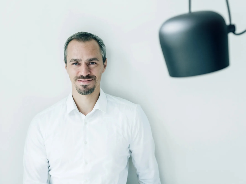 Ein Mann in weißem Hemd steht vor einer weißen Wand neben einer schwarzen Hängelampe.