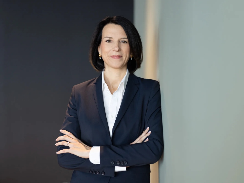 Eine Frau mit dunklem Haar in einem dunklen Blazer vor unscharfem Hintergrund.