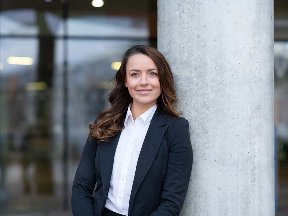A person in a black suit leaning against a grey column against a blurry background.