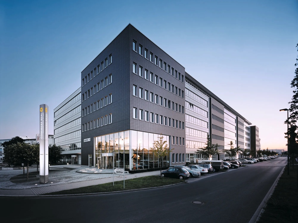 Modernes Bürogebäude mit Glasfassade bei Sonnenuntergang, umgeben von geparkten Autos und Bäumen.