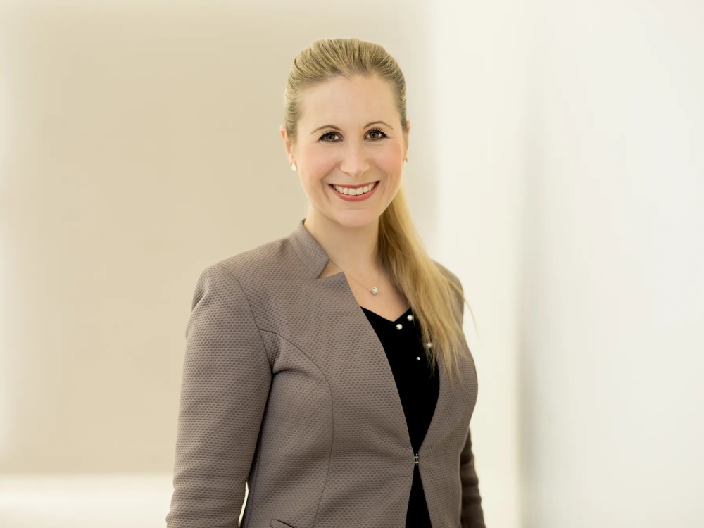 A smiling woman with a blonde ponytail in a grey blazer against a light background.