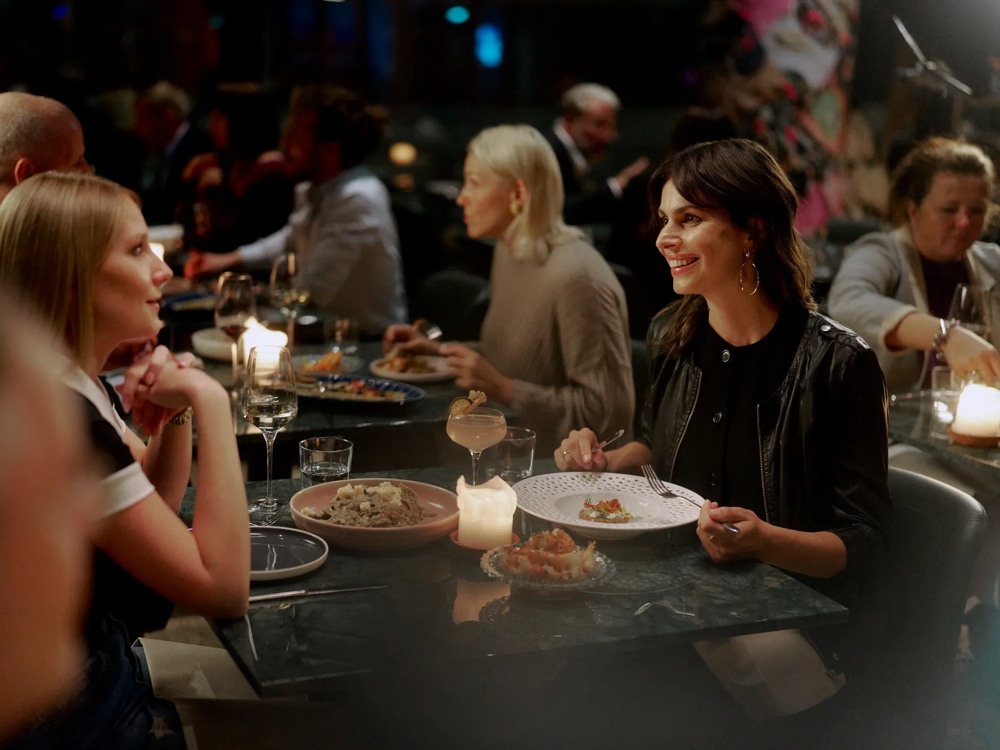 Zwei Frauen sitzen lächelnd bei Kerzenschein an einem Tisch in einem belebten Restaurant.