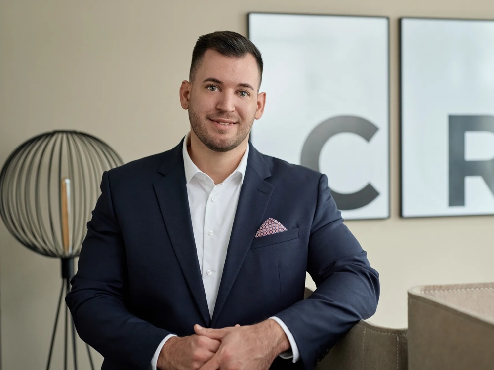 Ein Mann trägt einen schwarzen Anzug ohne Krawatte und ein Einstecktuch. Er steht mit einem Arm auf einer Abgrenzung gelehnt vor den gerahmten Initialen der Commerz Real und einem Lampenschirm.