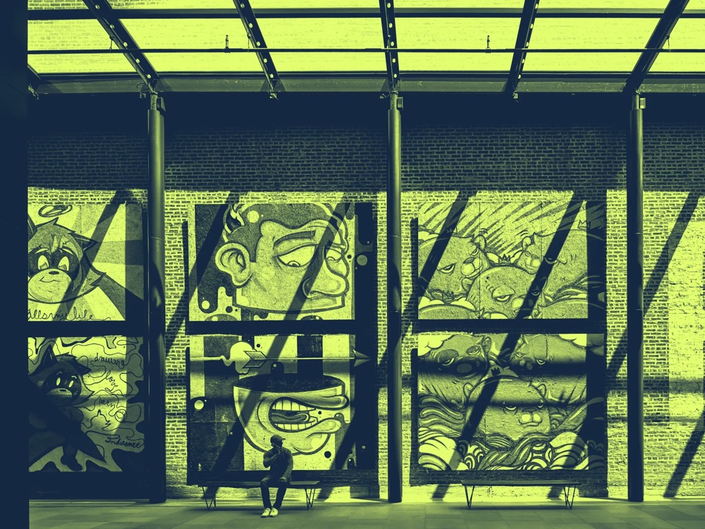 Wandgemälde mit Cartoon-Gesichtern in einer Halle, zwei Personen davor, Schatten von Glasdach fallen auf die Wand.