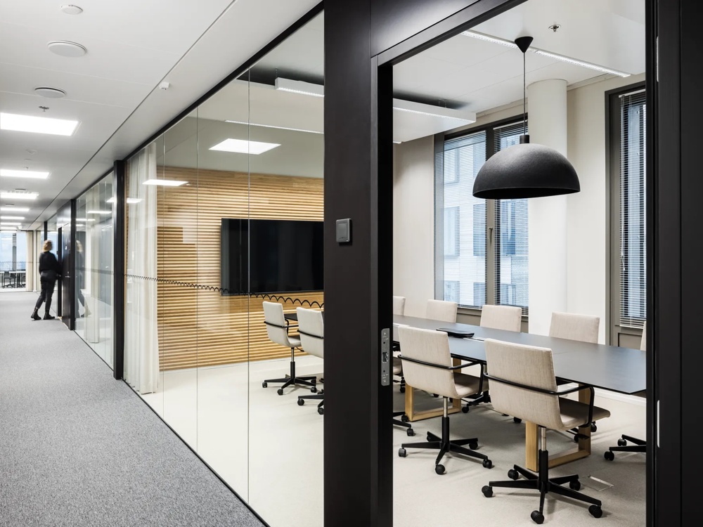 Moderner Büroflur mit Glaswänden, ein Konferenzraum mit Tisch und Stühlen, zwei Personen im Hintergrund.