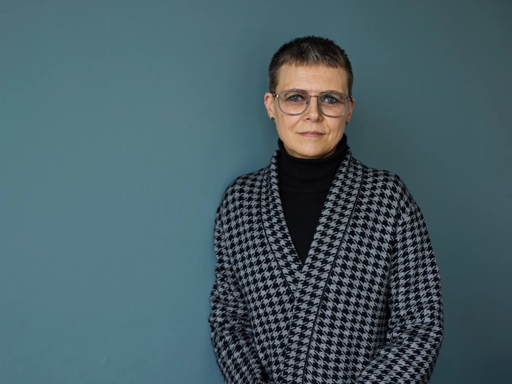 Eine Person mit Brille, kurzem Haar und kariertem Mantel vor blauem Hintergrund.