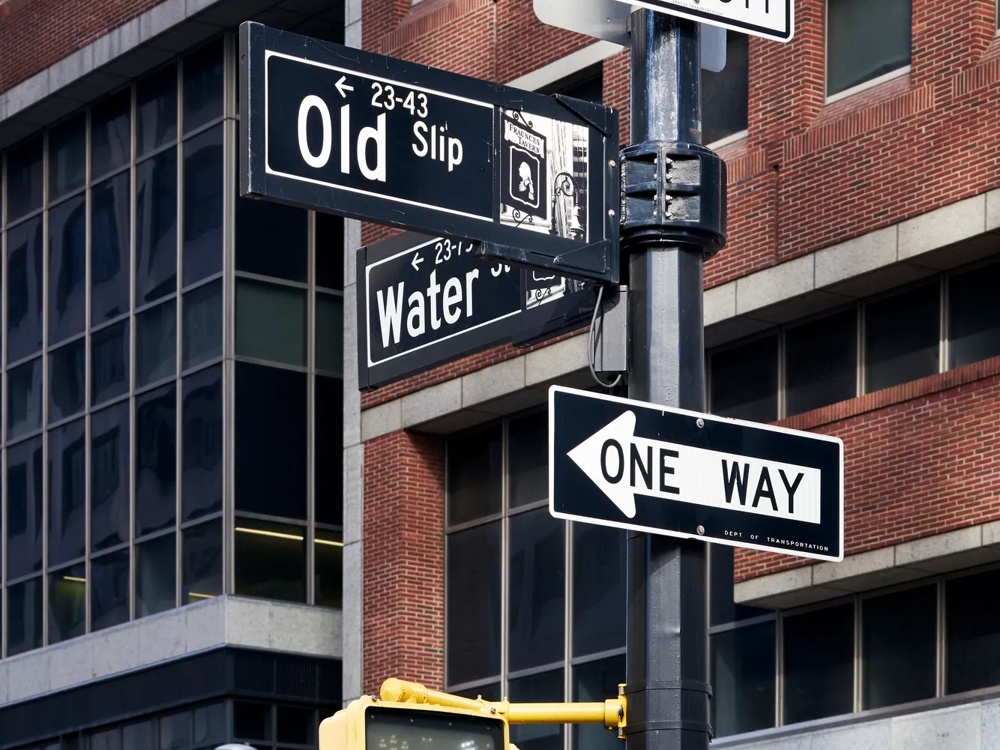 Road signs at an intersection in front of a brick building, including “One Way” and “No Left Turn”.