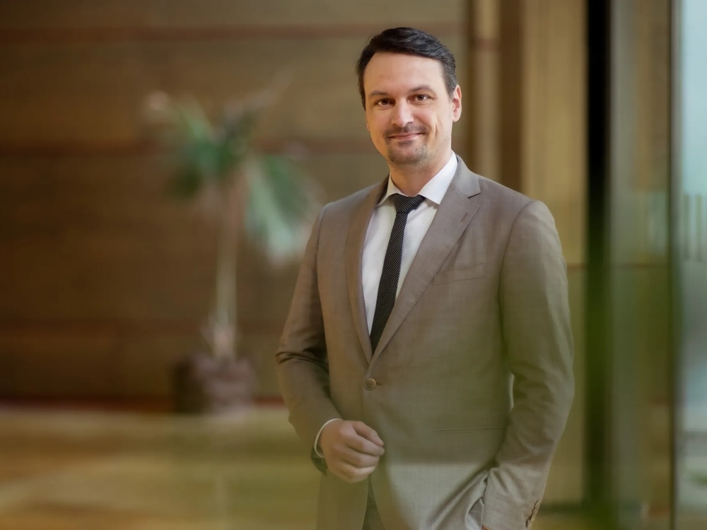 Ein Mann im Anzug lächelt in einem modernen Büro mit unscharfer Pflanze im Hintergrund.