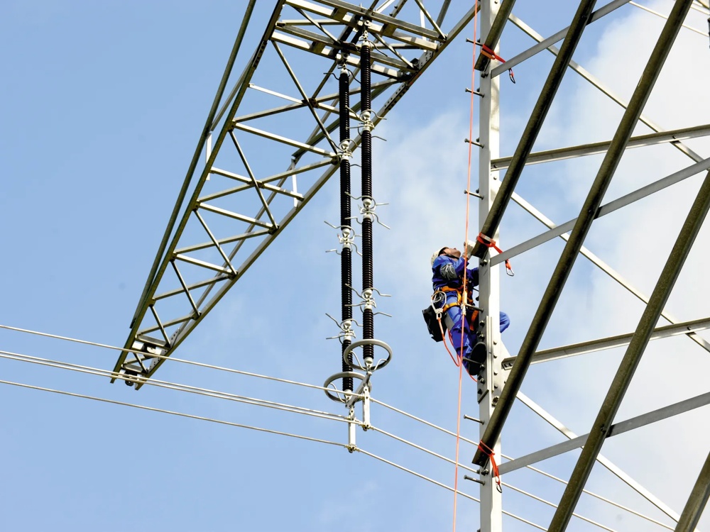 Ein Arbeiter in blauer Schutzkleidung klettert an einem Strommast bei klarem Himmel.