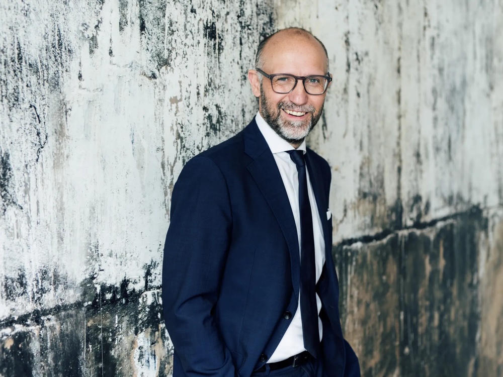 Ein Mann im Anzug steht lächelnd vor einer strukturierten, weißen Wand.