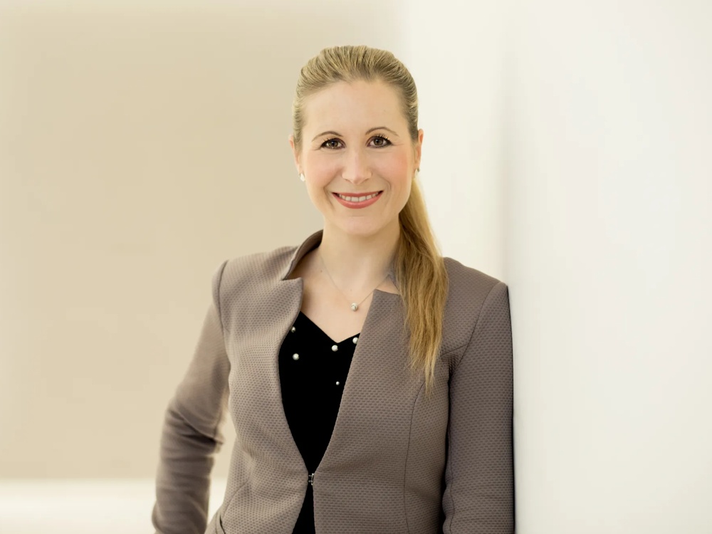 Woman with a blonde ponytail smiling into the camera, wearing a grey blazer, standing in front of a light background.