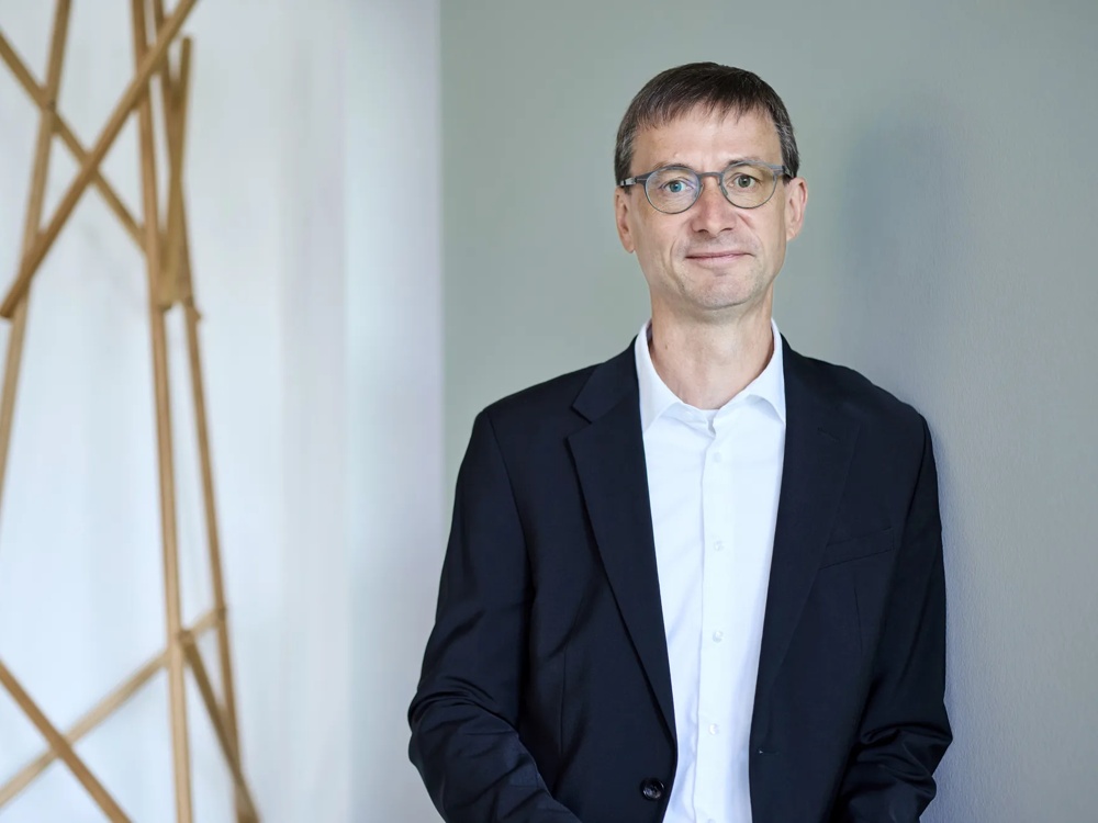 Ein Mann mit Brille im Anzug steht vor einer grauen Wand und schaut freundlich in die Kamera.