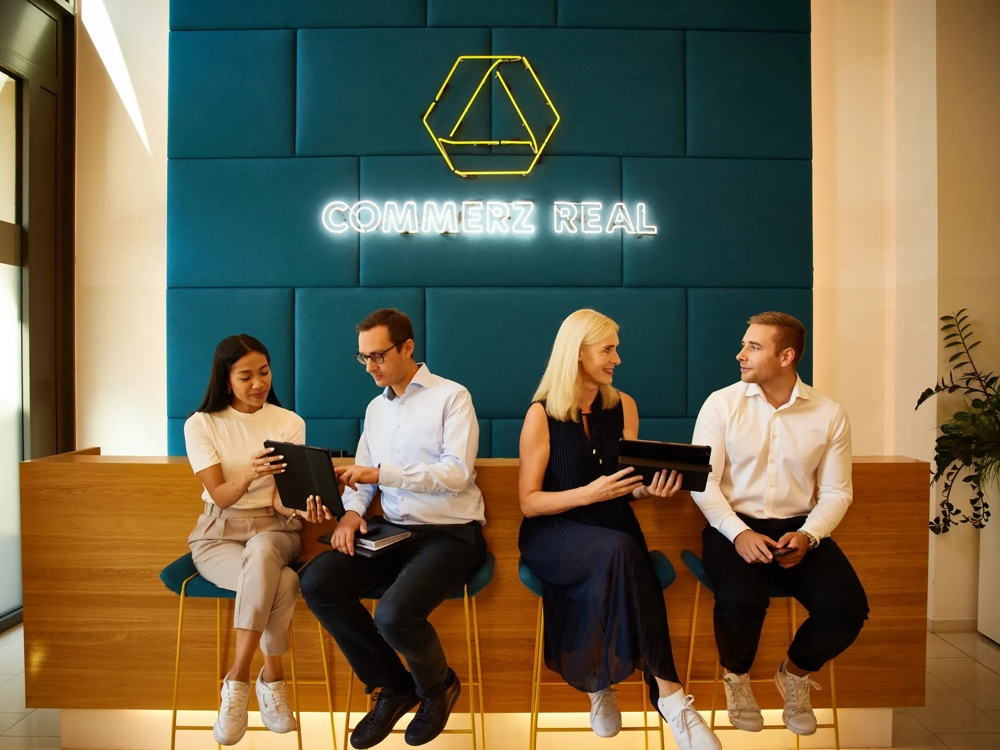 Vier Personen, darunter CEO Henning Koch, sitzen auf Barhockern vor einer Theke aus Holz, zwei von ihnen haben iPads in der Hand, die sie ihrem Nachbarn zeigen. Im Hintergrund ist eine petrolfarbene Wand auf der mit Neonschrift Commerz Real steht mit einem CR Logo aus gelber Neonschrift.