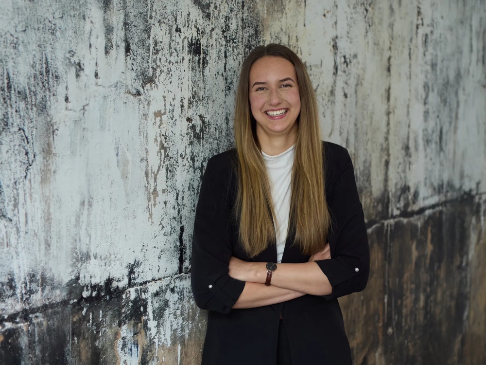 Commerz Real employee Marie Lysann Ulbricht, Key Account Manager for Sales Private and Business Clients NOM, is represented. She is standing in front of a weathered wall with her arms crossed, wearing a white shirt and smiling openly.