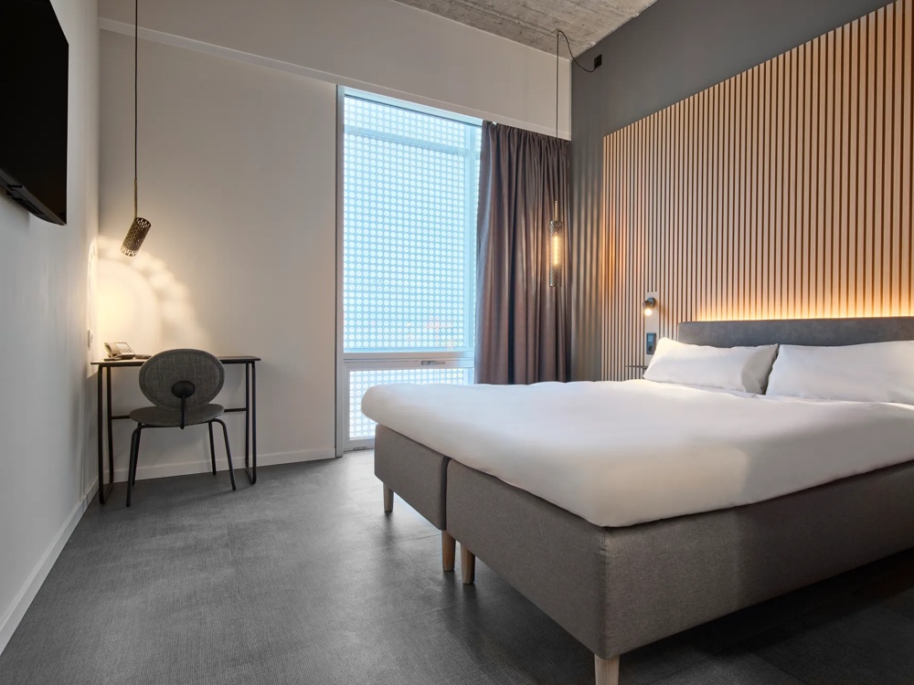 Modern hotel room with large bed, wooden panel wall, desk, chair and hanging lamp next to a window.