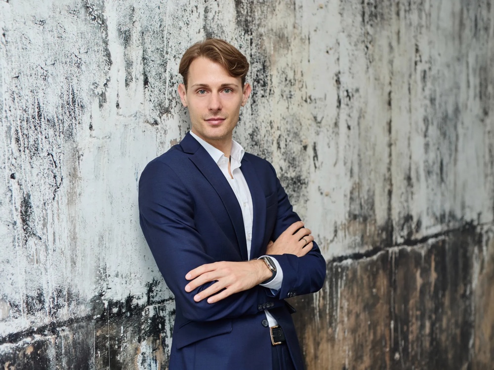 A man in a blue suit standing with arms crossed in front of a textured grey wall.