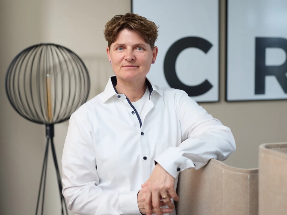 Eine Person in weißem Hemd lehnt an einem Stuhl, im Hintergrund eine Lampe und Buchstaben C, R.