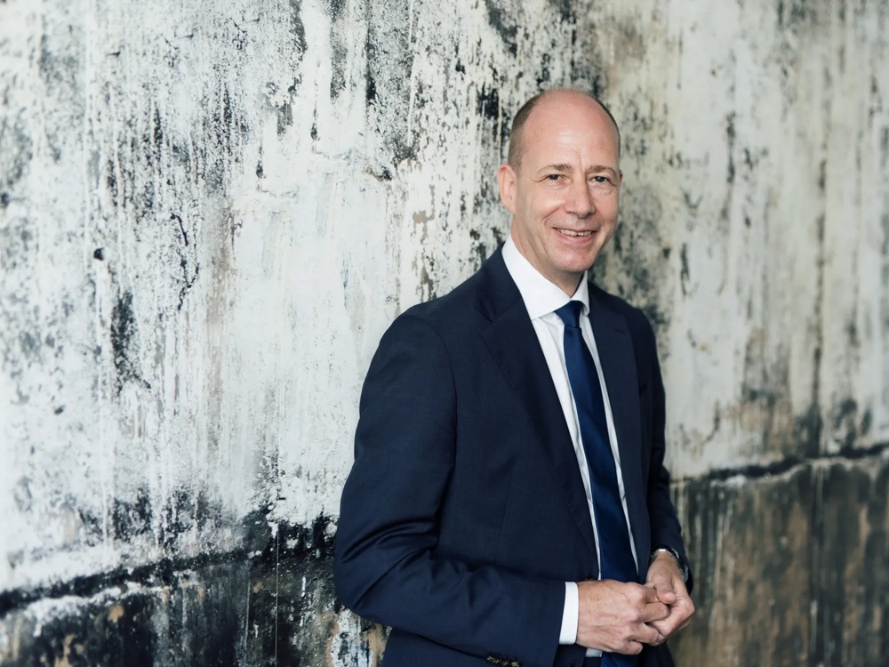 Ein Mann im Anzug steht vor einer strukturierten, verwitterten Wand, lächelt in die Kamera.