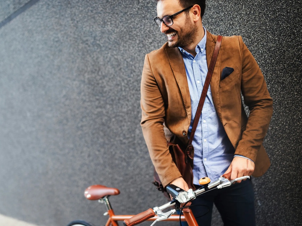 Mann in braunem Sakko mit Fahrrad vor grauer Wand, lächelnd.