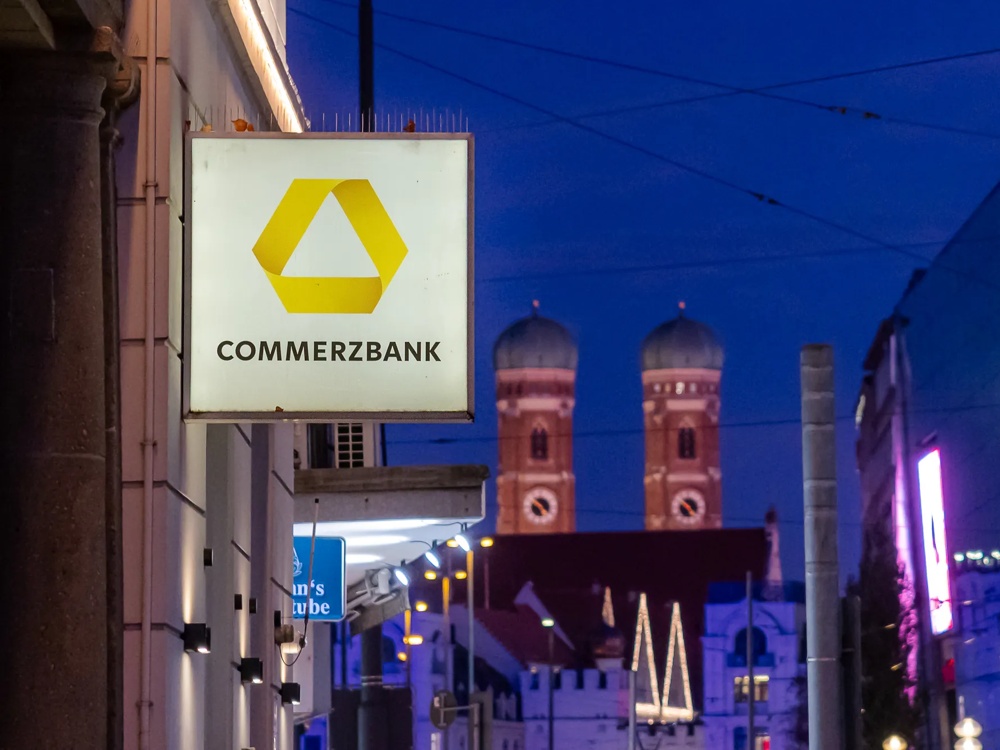 Häuserschlucht mit einer Hausfassade, an der ein Werbeschild einer Commerzbank Filiale hängt.