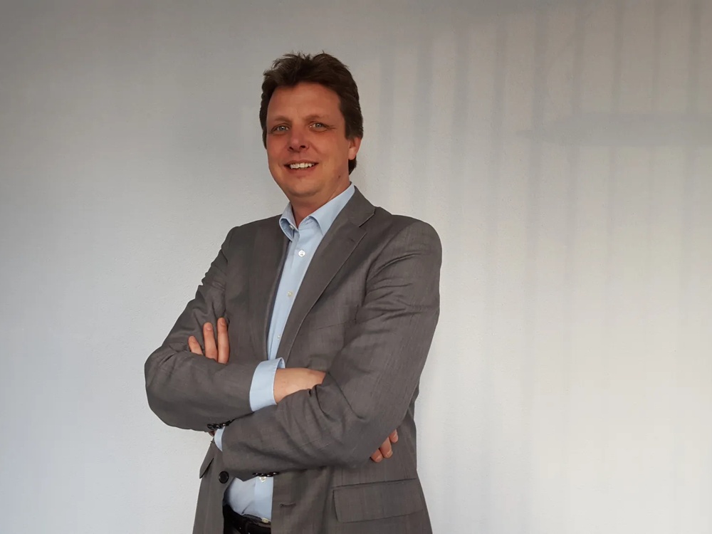 Ein Mann im grauen Anzug steht lächelnd mit verschränkten Armen vor einer weißen Wand.