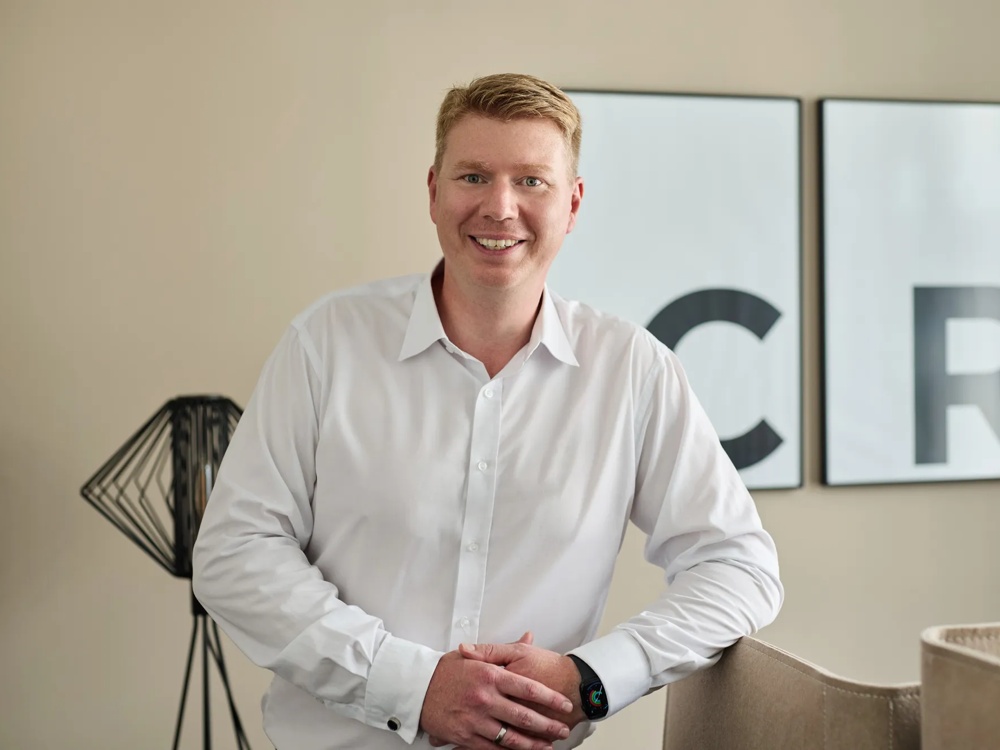 A smiling man in a white shirt leaning on a chair, with two large letters C and R in the background.