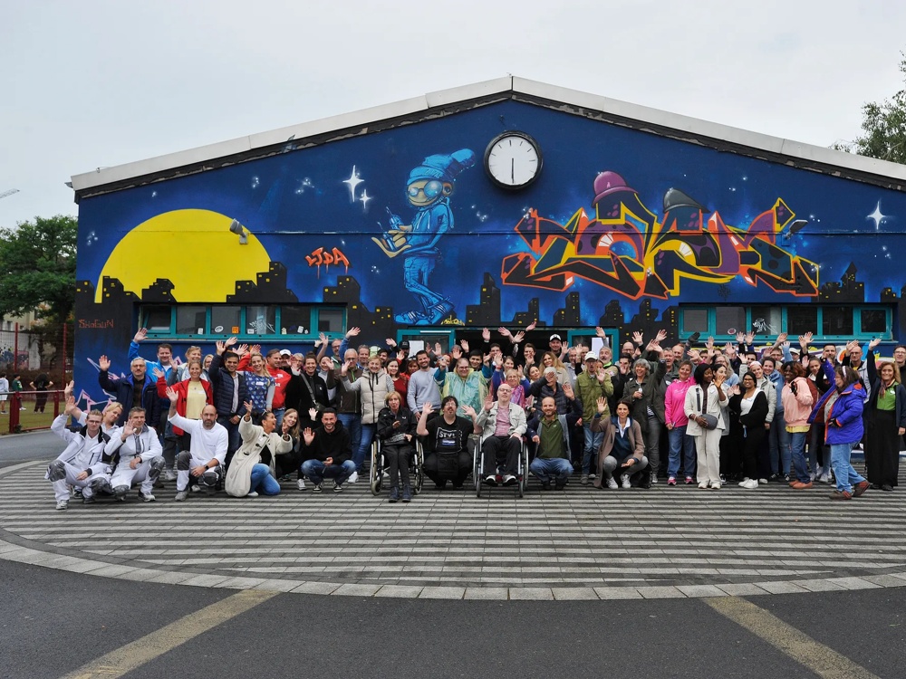 A large group of people posing in front of a building with colourful graffiti and a clock.