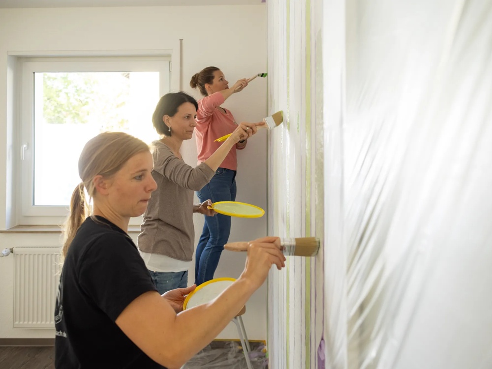 Drei Frauen streichen eine Wand mit Pinseln und gelben Farbschalen.