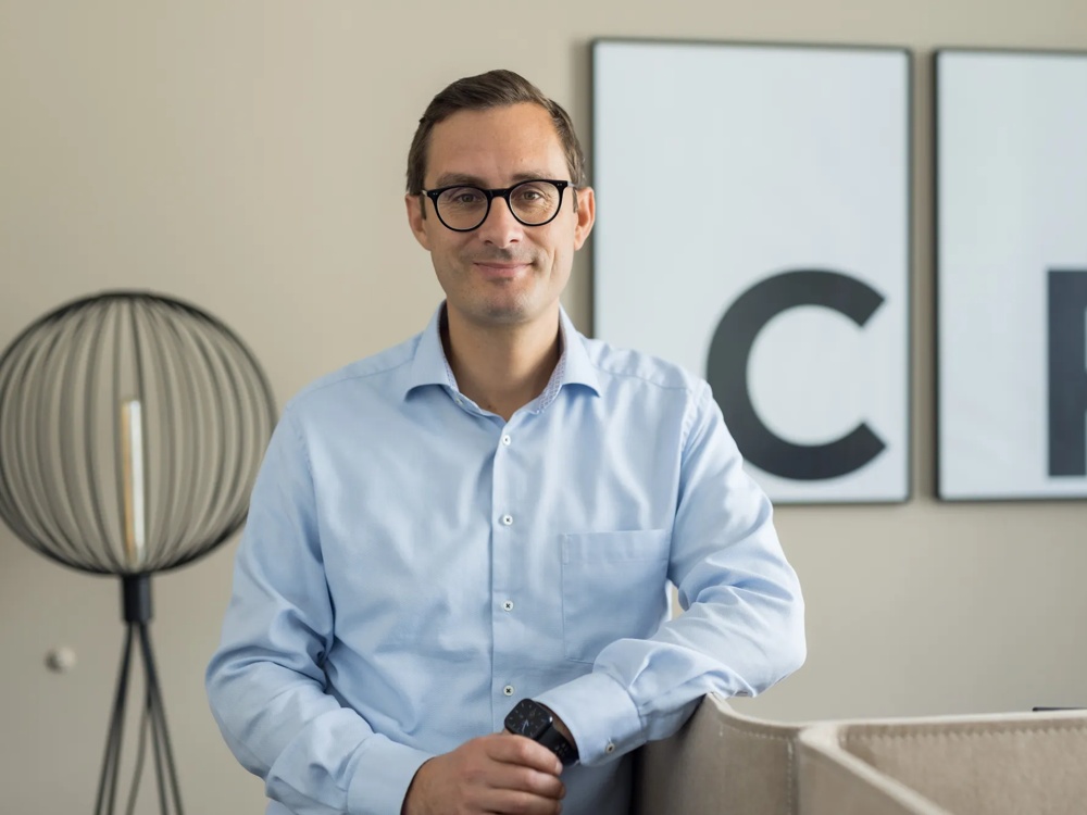 Ein Mann in blauem Hemd steht lächelnd vor zwei großen Buchstaben an der Wand.